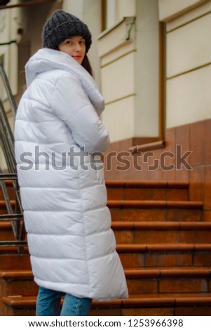 beautiful brunette in warm white coat or down jacket on a cold, gloomy street poses with a smile for a photo, an attractive woman in a stylish woolen coat in the winter on the street with a smile