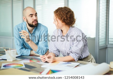 Content confident handsome creative designer leaning on table with color swatches and pencils and offering his idea to colleague at meeting in office