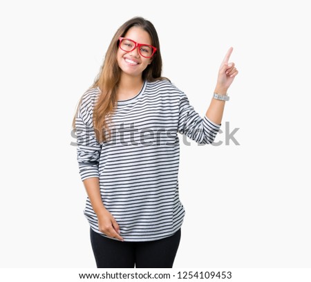 Young beautiful brunette woman wearing stripes sweater and red glasses over isolated background with a big smile on face, pointing with hand and finger to the side looking at the camera.