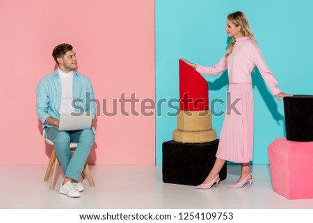 woman posing with nail polish and lipstick models while man sitting on chair and using laptop on pink and blue background