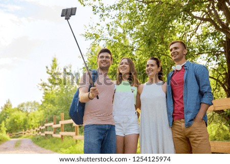 technology, friendship and hike concept - group of smiling friends with backpack taking picture by smartphone on selfie stick in summer