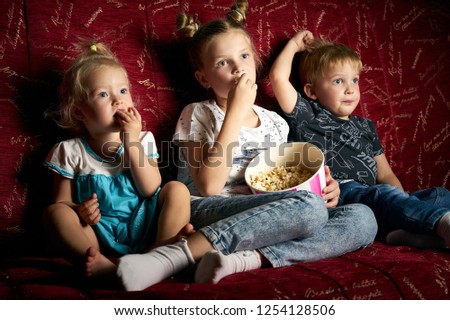 Children's movies: Three children watch movies at home on a big red sofa in the dark and eat popcorn.