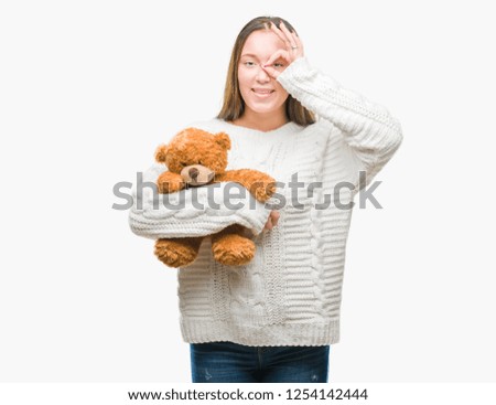 Young caucasian woman holding teddy bear over isolated background with happy face smiling doing ok sign with hand on eye looking through fingers