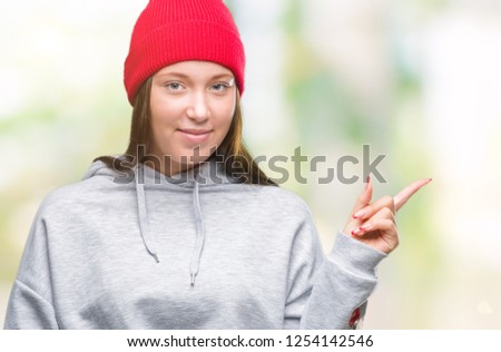 Young caucasian beautiful woman wearing wool cap over isolated background with a big smile on face, pointing with hand and finger to the side looking at the camera.