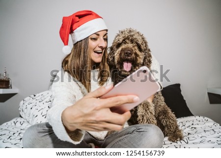 
Young and cheerful woman playing with her nice spanish water dog on Christmas morning, taking some pictures with her smart phone. Both with christmas costume. Lifestyle.