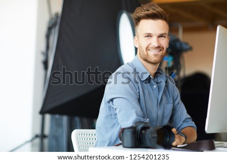 Portrait of handsome male photographer looking at camera while working in studio sitting at computer desk against lighting equipment, copy space