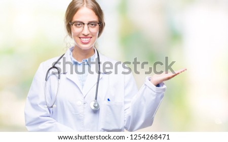 Beautiful young blonde doctor woman wearing medical uniform over isolated background smiling cheerful presenting and pointing with palm of hand looking at the camera.