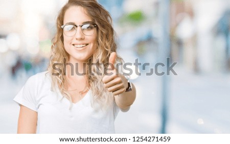 Beautiful young blonde woman wearing glasses over isolated background doing happy thumbs up gesture with hand. Approving expression looking at the camera showing success.