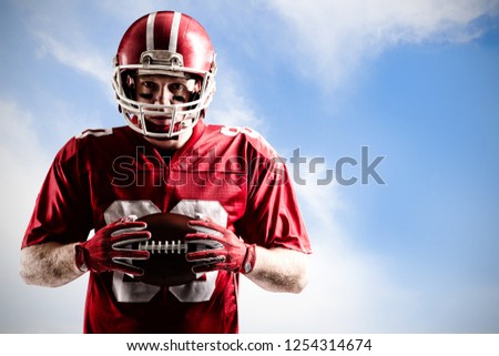 American football player standing with rugby helmet and ball against blue sky with clouds 