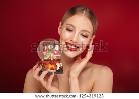 Beauty portrait of a cheerful pretty young topless woman standing isolated over red background, holding Christmas snow globe