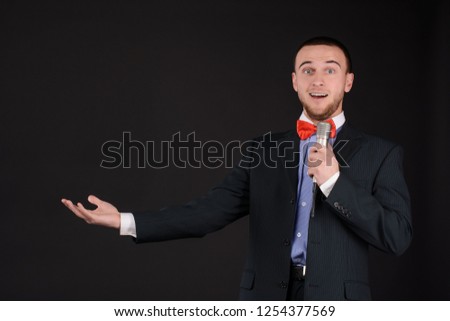 Surprised Handsome Master of ceremonies in black suit holding microphone in hand on black background. Showman, tv