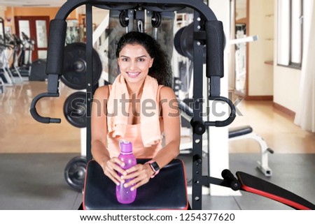 Picture of a happy woman holding a bottle of water while resting on a fitness machine. Shot in the gym center