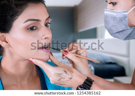 Picture of makeup artist using a makeup brush on her client lips with purple lipstick at home
