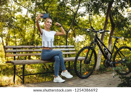 Image of athletic woman sitting on bench in park with bicycle and taking selfie photo on cell phone