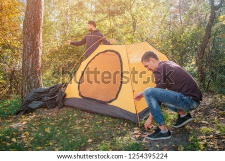 Picture of two young men in forest putting tent. One guy works with ropes. Another stand behind tent. They work together