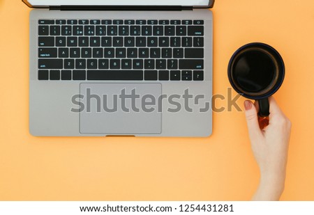 Hand holds a cup of coffee and a laptop on an orange background, a view from above, flatlay. Work on a laptop is isolated on a color background.
