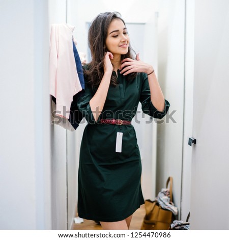 Portrait of a young and beautiful Indian Asian woman trying out a green dress in a fitting room in a fashion shop in a mall. She is smiling happily as she tries on her new clothing.