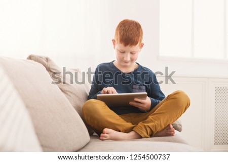 Boy playing online games on digital tablet on sofa in light room