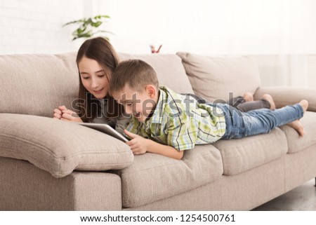 Happy siblings watching cartoons on tablet together lying on sofa at home
