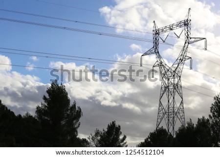 Outdoor view of the silhouette of a high metallic electrical pylon with a blue sky in background and white clouds. Symbolic picture of transmission of energy. Pattern of cables and wires. Top of trees
