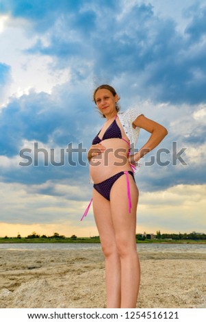 A happy and joyful pregnant woman stands by the sea and ocean on the beach under the sun in the open air, hair develops in the wind. Wide-angle photo.
