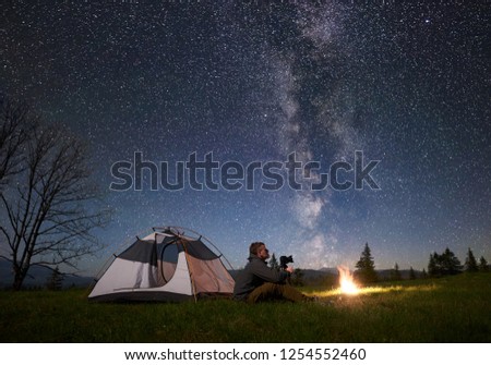 Beautiful evening in mountains. Young man with photo camera sitting alone in front of tourist tent at burning bonfire on grassy valley under dark blue starry sky. Tourism and recreation concept.