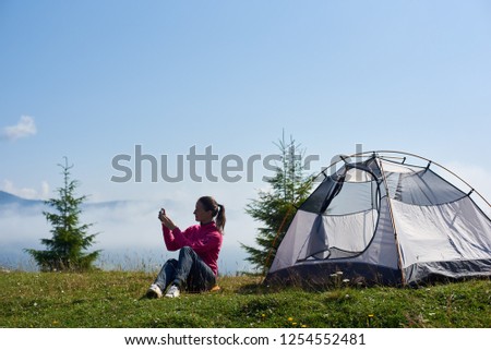 Young tourist girl sitting on green grass in front of small tourist tent and taking picture of beautiful foggy mountains covered with white clouds under clear blue sky on bright summer morning.
