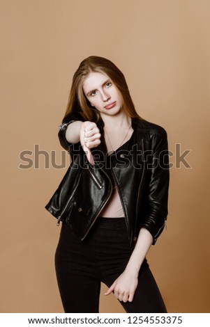 Human emotion. Young pretty woman in leather jacket posing on yellow beige background. Blonde girl with long hair portrays sadness, she is upset and shows finger down