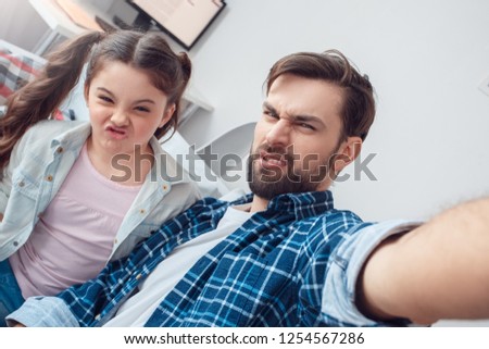 Bearded man and little girl at home family time sitting on sofa taking selfie pictures grimace disgusted close-up