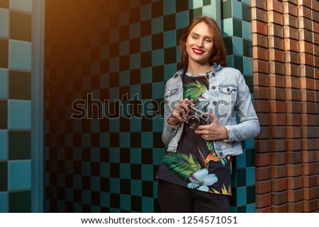 Cool funny girl model with retro film camera wearing a denim jacket, dark hair outdoors over city wall in a cage background. Sun flare