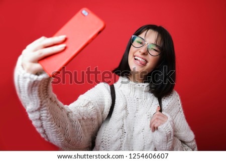 Charming modern woman in sweater and glasses taking selfie and showing tongue out on red backdrop