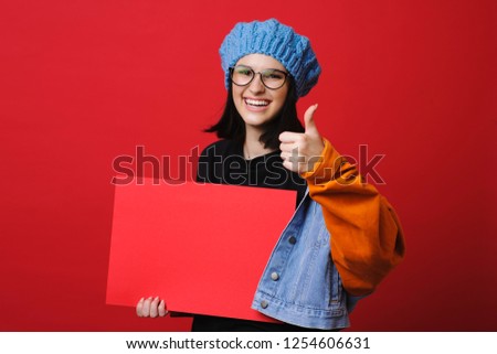 Pretty brunette in jacket holding thumb up while showing blank red paper smiling at camera