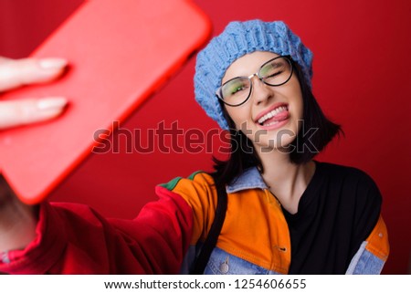 Pretty playful woman in ht and colorful jacket showing tongue and taking selfie on red background