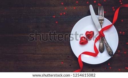 Valentine's Day Festive table setting, flat lay with two red heart shape chocolate candies on white plate, fork, knife and red ribbons on wooden table. Valentine Day, love, dating concept, copy space Royalty-Free Stock Photo #1254615082