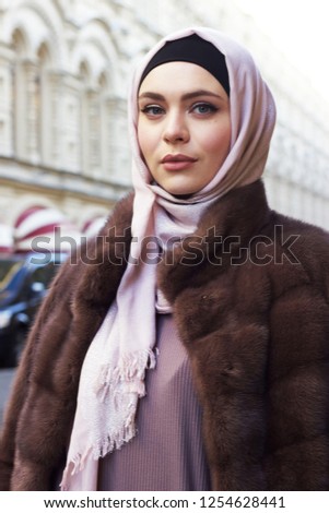An adorable Muslim girl in rose hijab, long dress and fur on the street of a big city Royalty-Free Stock Photo #1254628441