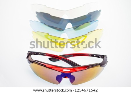 Stylish cycling sports glasses with red frames in the foreground, close-up. In the background, a set of different colors of spare glasses for sports glasses. Background white blurred