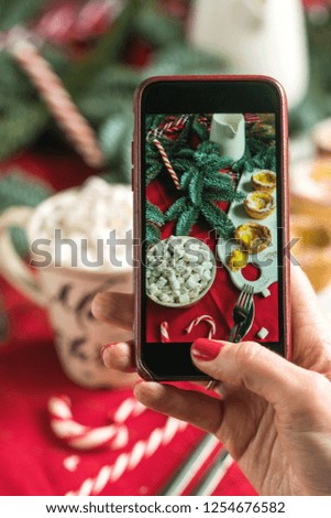 The hands of the girl with the phone make a photo of the New Year mood. Cocoa, marshmallows, cup, spruce, red cloth.