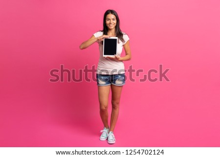 Full length portrait of a satisfied pretty girl pointing finger at blank screen tablet computer isolated over pink background