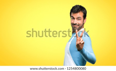 Man wearing an apron smiling and showing victory sign on yellow background
