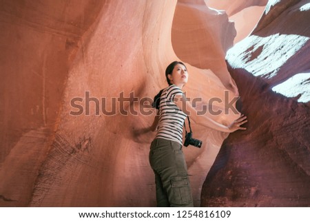 young female traveler touching the rock turning back her head while hiking in upper antelope canyon. tourist with camera carefully climbing the desert cave on sunny day. asian girl love travel wild.