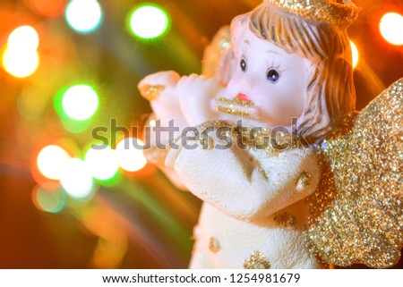 Figurine of little angel with golden shiny wings and halo that plays flute on shining background of blurry flashes. Christmas or New Year picture of cute angel sculpture on bokeh lights background