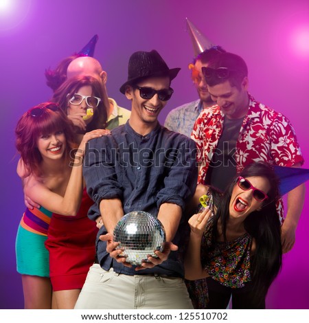 young handsome caucasian man holding a disco ball in his hands and looking at it  with other happy people around him with party horns and hats laughing