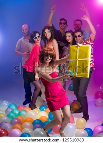 young attractive caucasian girl holding a gift box in her hands and laughing, with other people having fun in the background surounded with colorful balloons and lights
