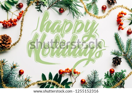 flat lay with festive arrangement of pine tree branches, common sea buckthorn and christmas decorations on white tabletop with "happy holidays" lettering