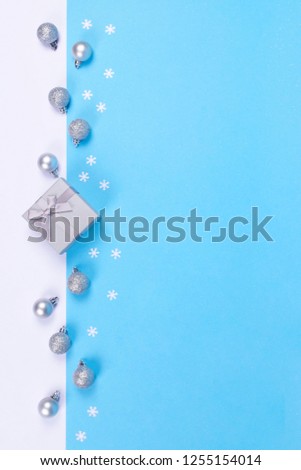 Beautiful gift box tied with little bow and silver glittering and shiny Christmas baubles on white and blue background decorated with snowflake confetti.