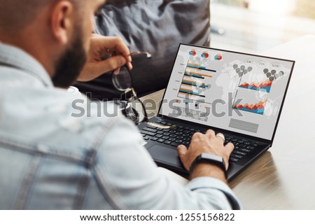 Back view. Young male hipster freelancer sits in cafe at table and works on laptop computer with graphs, charts, diagrams on monitor.View of computer screen and male hand on keyboard. Online education Royalty-Free Stock Photo #1255156822