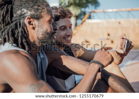Two men looking at the mobile phone while sitting on an outdoor basketball court.