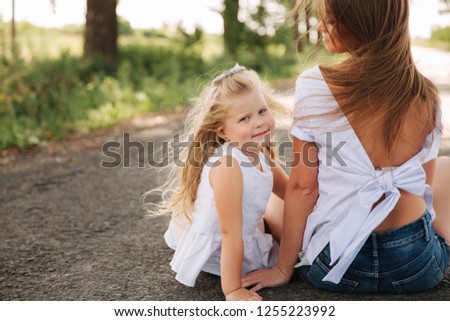 Attractive Mom and blonde hair daughter sits on road near big alley. They smile and look to natune. Front view