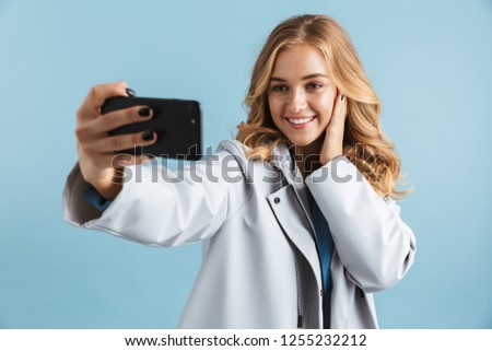 Cheerful young girl wearing raincoat standing isolated over blue background, taking a selfie