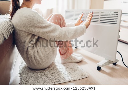 Using heater at home in winter. Woman warming her hands sitting by device and wearing warm clothes. Heating season. Royalty-Free Stock Photo #1255238491